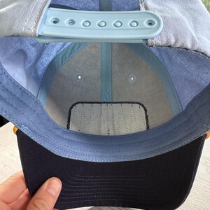 May include: A light blue baseball cap with a dark navy blue brim. The interior features a light blue plastic snapback closure, light blue fabric lining, and a white rectangular patch on the front panel. The cap is constructed with six panels.