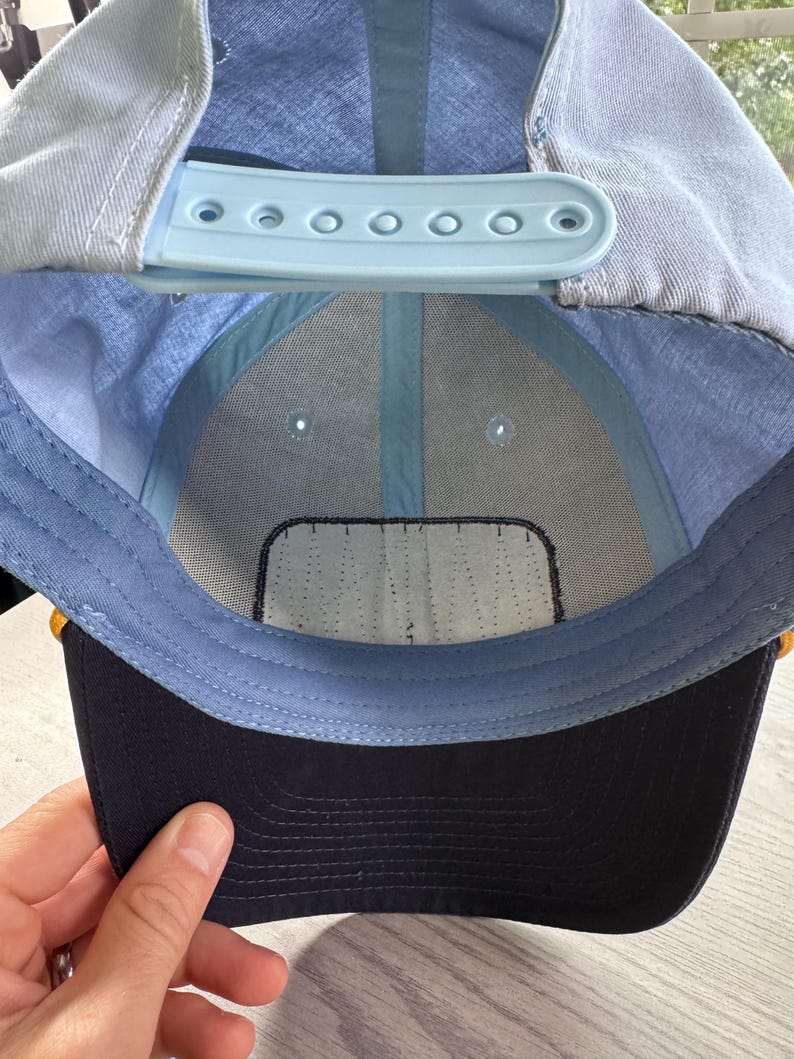 May include: A light blue baseball cap with a dark navy blue brim. The interior features a light blue plastic snapback closure, light blue fabric lining, and a white rectangular patch on the front panel. The cap is constructed with six panels.