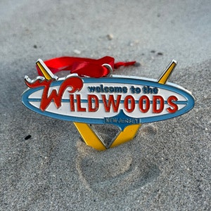May include: A silver metal ornament shaped like a surfboard with the words "Welcome to the Wildwoods, New Jersey" in red and blue lettering.