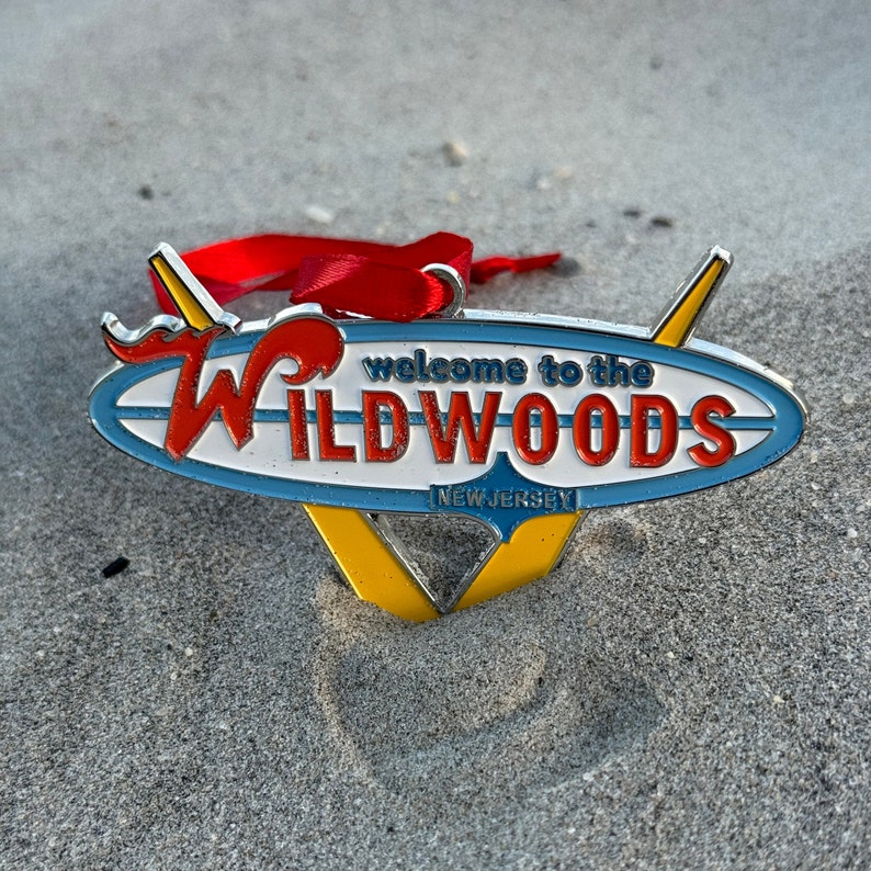 May include: A silver metal ornament shaped like a surfboard with the words "Welcome to the Wildwoods, New Jersey" in red and blue lettering.