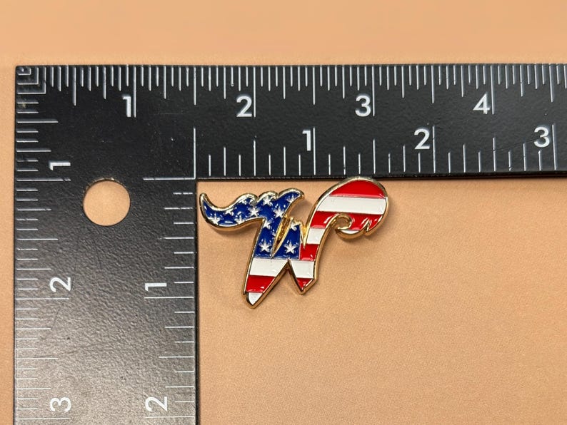 May include: A gold-colored metal pin in the shape of the letter "W". The pin is painted with the American flag design, featuring red and white stripes and blue with white stars. The pin is approximately 1 inch in length.