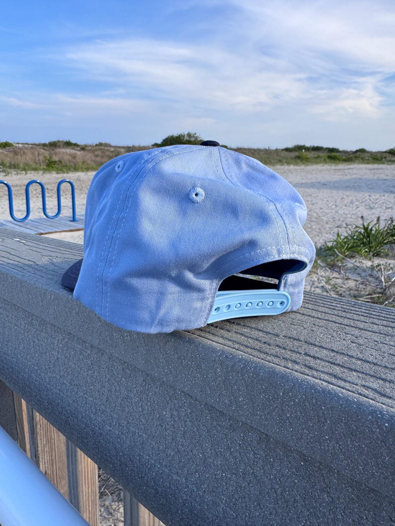 May include: A light blue baseball cap with a dark blue brim and a matching light blue plastic snapback closure. The cap features a structured crown with stitched eyelets and a dark blue button at the top, showcasing a classic six-panel design.