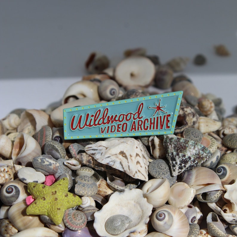 May include: A blue and red enamel pin with the text "Wildwood Video Archive" and a starburst design. The pin is surrounded by a variety of seashells.