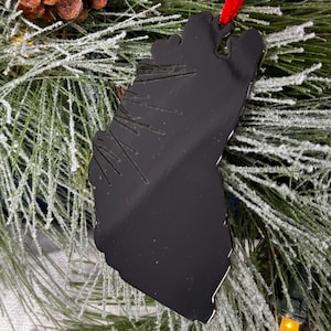 May include: Black metal Christmas ornament shaped like a tree, hanging on a frosted pine branch with a red ribbon.