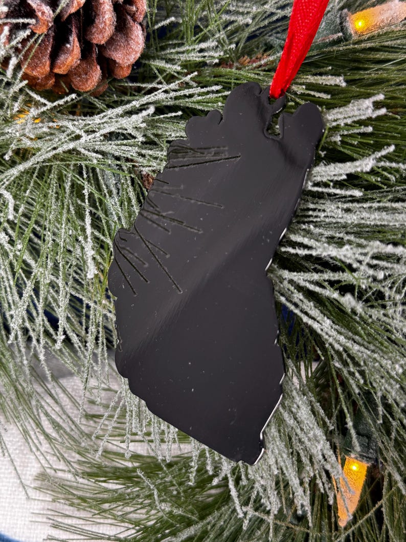 May include: Black metal Christmas ornament shaped like a tree, hanging on a frosted pine branch with a red ribbon.