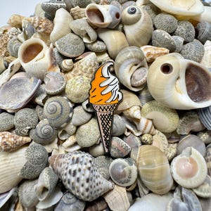 Op de afbeelding: Een close-up van een oranje en witte emaille pin in de vorm van een ijshoorn. De pin is omringd door verschillende schelpen.