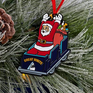 May include: A red and white Santa Claus ornament riding a blue and white roller coaster with the words "Great White" on the side. The roller coaster has a shark fin on the front.