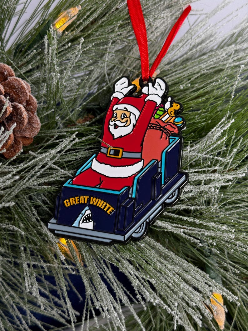 May include: A red and white Santa Claus ornament riding a blue and white roller coaster with the words "Great White" on the side. The roller coaster has a shark fin on the front.