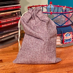 May include: A small, gray burlap sack with a drawstring closure. The sack is sitting on a wooden surface.