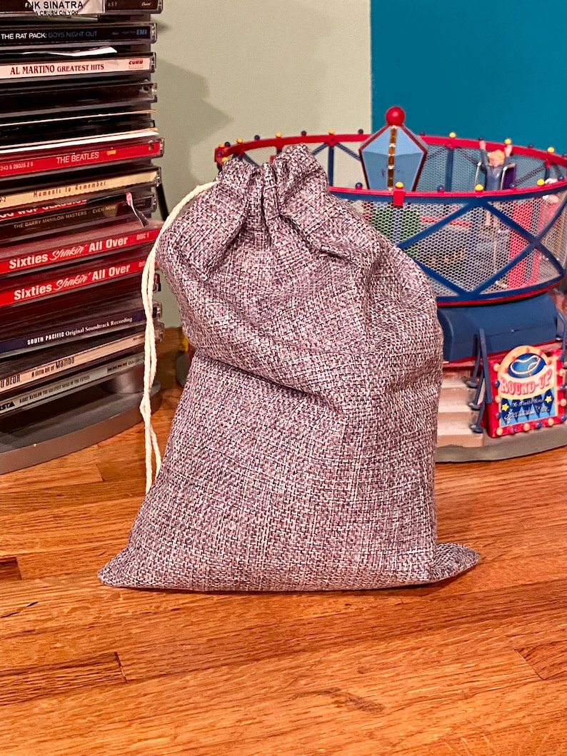 May include: A small, gray burlap sack with a drawstring closure. The sack is sitting on a wooden surface.