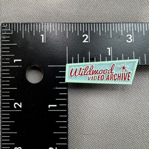 May include: A light blue enamel pin with a red and white sign that reads "Wildwood Video Archive" with a yellow starburst and a red star.