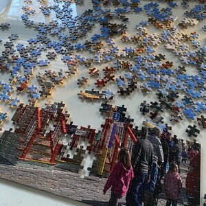 May include: A partially completed jigsaw puzzle of a boardwalk scene with people walking on the boardwalk. The puzzle is set on a white surface. The puzzle pieces are mostly blue and brown. The puzzle is missing some pieces. The puzzle is in the process of being assembled. The puzzle is a popular activity for people of all ages.