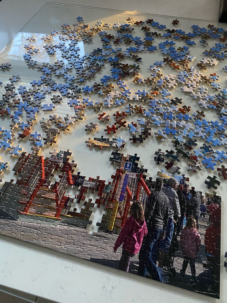 May include: A partially completed jigsaw puzzle of a boardwalk scene with people walking on the boardwalk. The puzzle is set on a white surface. The puzzle pieces are mostly blue and brown. The puzzle is missing some pieces. The puzzle is in the process of being assembled. The puzzle is a popular activity for people of all ages.