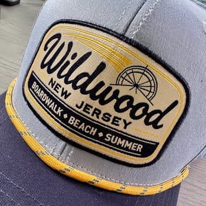 May include: A grey baseball cap featuring a rectangular patch with a tan background and black text that reads Wildwood New Jersey Boardwalk Beach Summer. The patch includes a graphic of a Ferris wheel and a yellow rope trim along the brim.