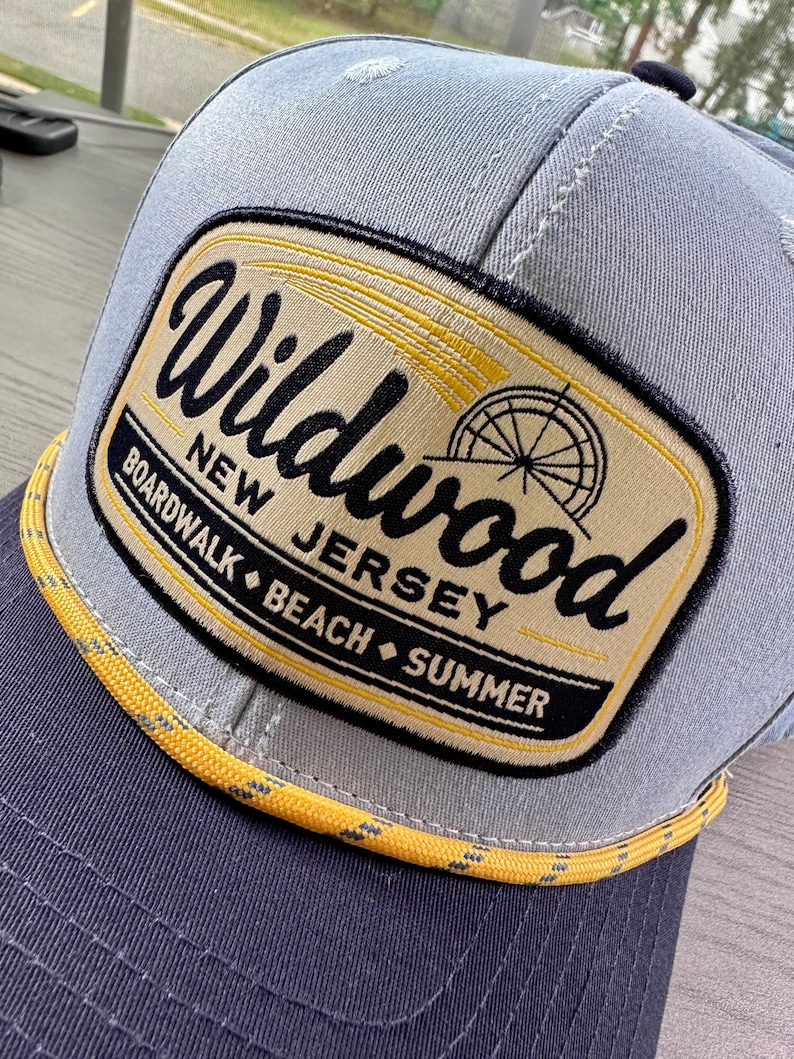 May include: A grey baseball cap featuring a rectangular patch with a tan background and black text that reads Wildwood New Jersey Boardwalk Beach Summer. The patch includes a graphic of a Ferris wheel and a yellow rope trim along the brim.
