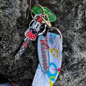 May include: A white and blue lanyard with a retro design featuring a silver metal charm that says "Wildwood". A Mickey Mouse key chain with red polka dots and a green hotel key are attached to the lanyard.