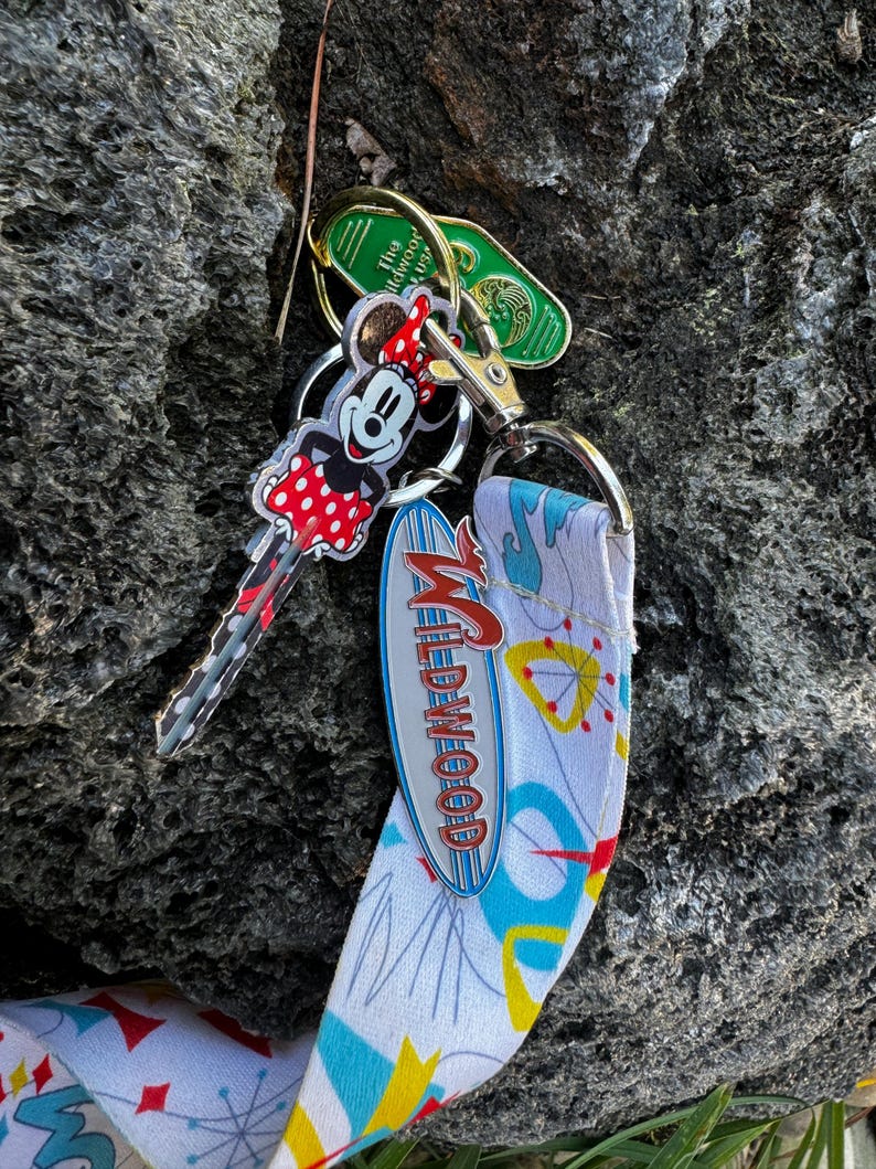 May include: A white and blue lanyard with a retro design featuring a silver metal charm that says "Wildwood". A Mickey Mouse key chain with red polka dots and a green hotel key are attached to the lanyard.