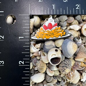 K&ouml;nnte beinhalten: Eine gelbe und orangefarbene Emaille-Anstecknadel mit einer Platte Spaghetti, die mit Schlagsahne und roten Erdbeeren belegt ist. Die Anstecknadel ist von Muscheln umgeben.