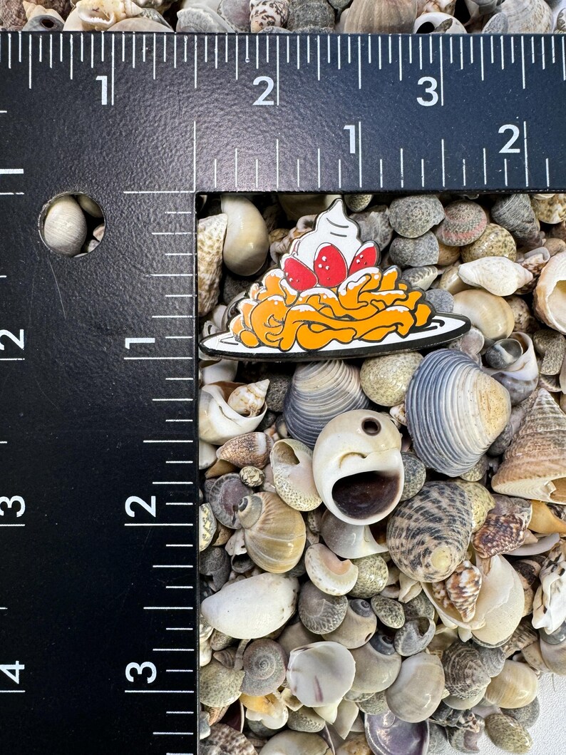 K&ouml;nnte beinhalten: Eine gelbe und orangefarbene Emaille-Anstecknadel mit einer Platte Spaghetti, die mit Schlagsahne und roten Erdbeeren belegt ist. Die Anstecknadel ist von Muscheln umgeben.