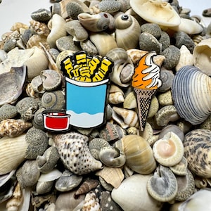 Op de afbeelding: Twee emaillepins op een bed van schelpen. E&eacute;n pin is een blauwe en witte cartoon van een bakje friet met een rood ketchupbakje. De andere pin is een cartoon van een oranje ijshoorn.
