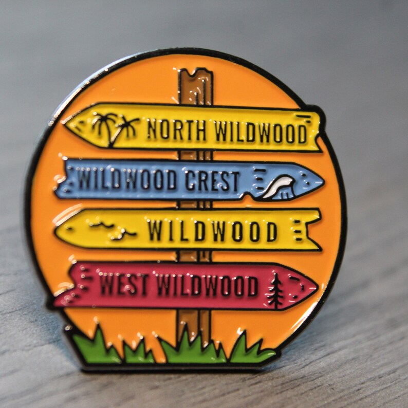 Pu&ograve; includere: Una spilla smaltata colorata che presenta un cartello di legno con quattro frecce direzionali che puntano a diverse localit&agrave;: North Wildwood, Wildwood Crest, Wildwood e West Wildwood. Il cartello &egrave; posizionato su uno sfondo arancione brillante con erba verde nella parte inferiore.