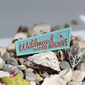 May include: A blue and yellow enamel pin with the text "Wildwood Video Archive" in red lettering and a red starburst design.