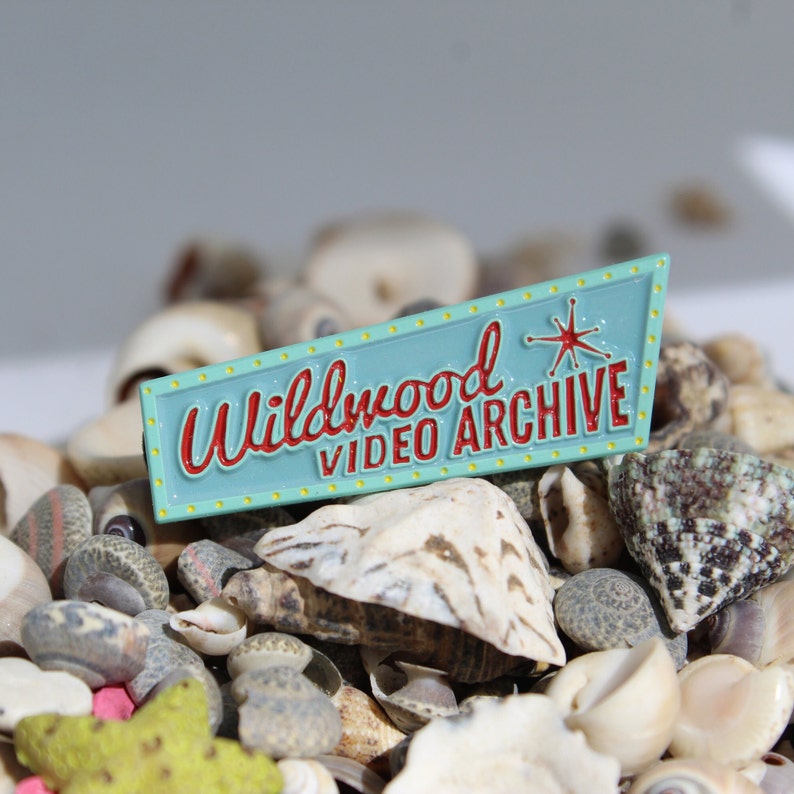 May include: A blue and yellow enamel pin with the text "Wildwood Video Archive" in red lettering and a red starburst design.