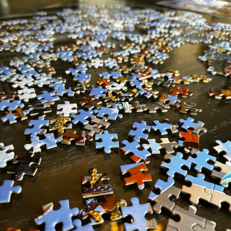 May include: A close-up of a partially completed jigsaw puzzle with blue puzzle pieces on a dark brown surface. The puzzle pieces are scattered across the surface, with some pieces overlapping.