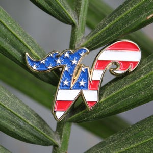 May include: A patriotic enamel pin in the shape of the letter "W", featuring the American flag design. The pin has a gold border and is set against a backdrop of green leaves. The pin's design incorporates red and white stripes, blue with white stars.