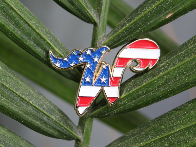 May include: A patriotic enamel pin in the shape of the letter "W", featuring the American flag design. The pin has a gold border and is set against a backdrop of green leaves. The pin's design incorporates red and white stripes, blue with white stars.