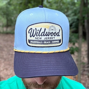 May include: A light blue baseball cap with a dark navy blue brim and a yellow rope detail. The front features a rectangular patch with the text Wildwood New Jersey Boardwalk Beach Summer and a graphic of a Ferris wheel in black and yellow.