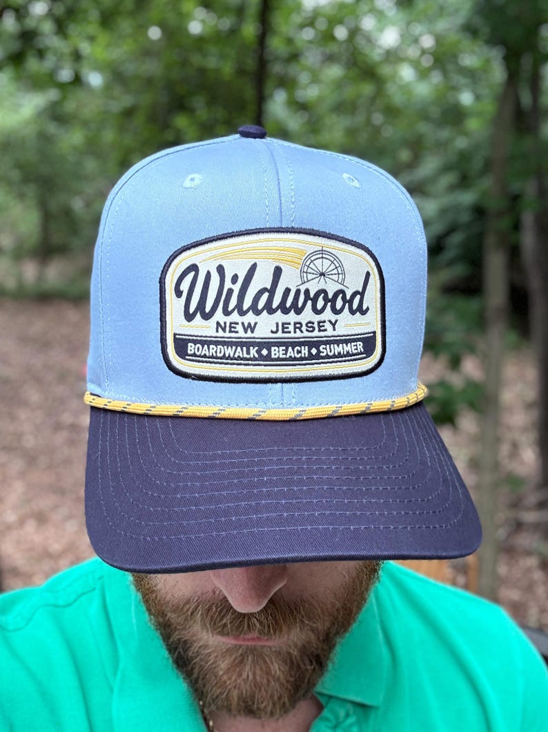 May include: A light blue baseball cap with a dark navy blue brim and a yellow rope detail. The front features a rectangular patch with the text Wildwood New Jersey Boardwalk Beach Summer and a graphic of a Ferris wheel in black and yellow.