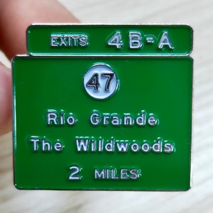 Puede incluir: Un pin de esmalte verde y blanco con un dise&ntilde;o de se&ntilde;al de tr&aacute;fico. El letrero dice "EXITS 4 B-A" en la parte superior, con "47" en un c&iacute;rculo blanco, y "Rio Grande, The Wildwoods 2 MILES" debajo.