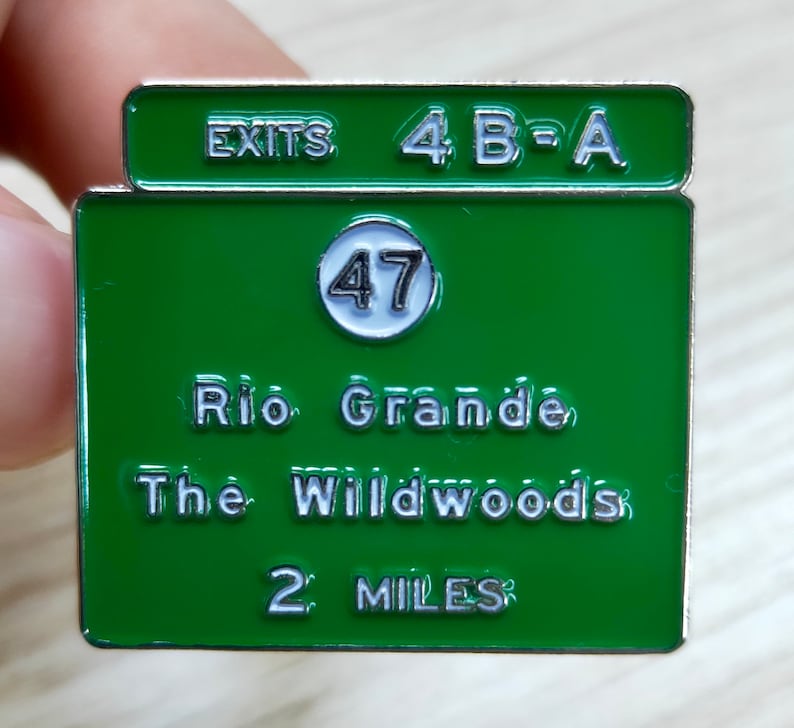 Puede incluir: Un pin de esmalte verde y blanco con un dise&ntilde;o de se&ntilde;al de tr&aacute;fico. El letrero dice "EXITS 4 B-A" en la parte superior, con "47" en un c&iacute;rculo blanco, y "Rio Grande, The Wildwoods 2 MILES" debajo.