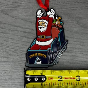 May include: A red and white Santa Claus ornament riding a blue and white roller coaster with the words "Great White" on the front. The ornament is about 2 inches tall and is hanging from a red ribbon.