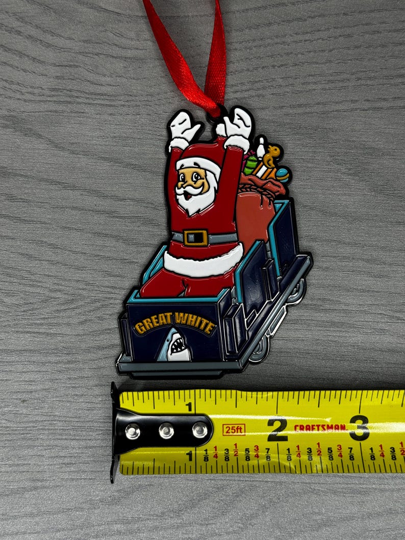 May include: A red and white Santa Claus ornament riding a blue and white roller coaster with the words "Great White" on the front. The ornament is about 2 inches tall and is hanging from a red ribbon.