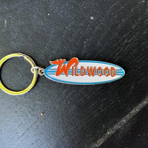 May include: A silver keychain with a yellow key ring and a blue and white surfboard charm with the word "WILDWOOD" in red letters.
