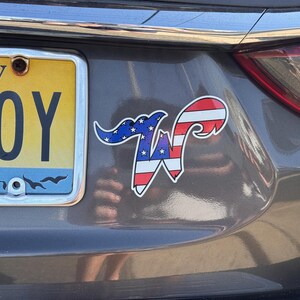 May include: A sticker on a car's rear with a patriotic design. The sticker features a stylized "W" with a blue section of stars and red and white stripes, resembling the American flag. The car has a New Jersey license plate.