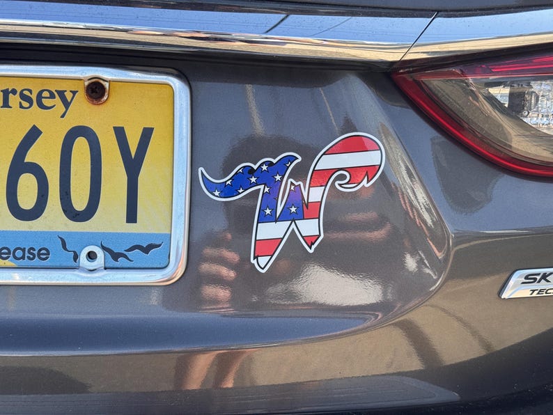 May include: A sticker on a car's rear with a patriotic design. The sticker features a stylized "W" with a blue section of stars and red and white stripes, resembling the American flag. The car has a New Jersey license plate.