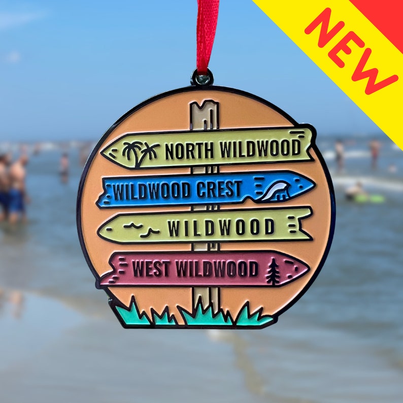 May include: A colorful metal signpost ornament with four directional signs. The signs read "North Wildwood", "Wildwood Crest", "Wildwood", and "West Wildwood". The ornament is decorated with palm trees, waves, and a pine tree.