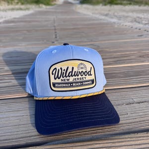 May include: A light blue baseball cap with a dark navy blue brim and a yellow rope detail. The front features a patch with the text Wildwood New Jersey Boardwalk Beach Summer and a graphic of a Ferris wheel in black, yellow, and white.