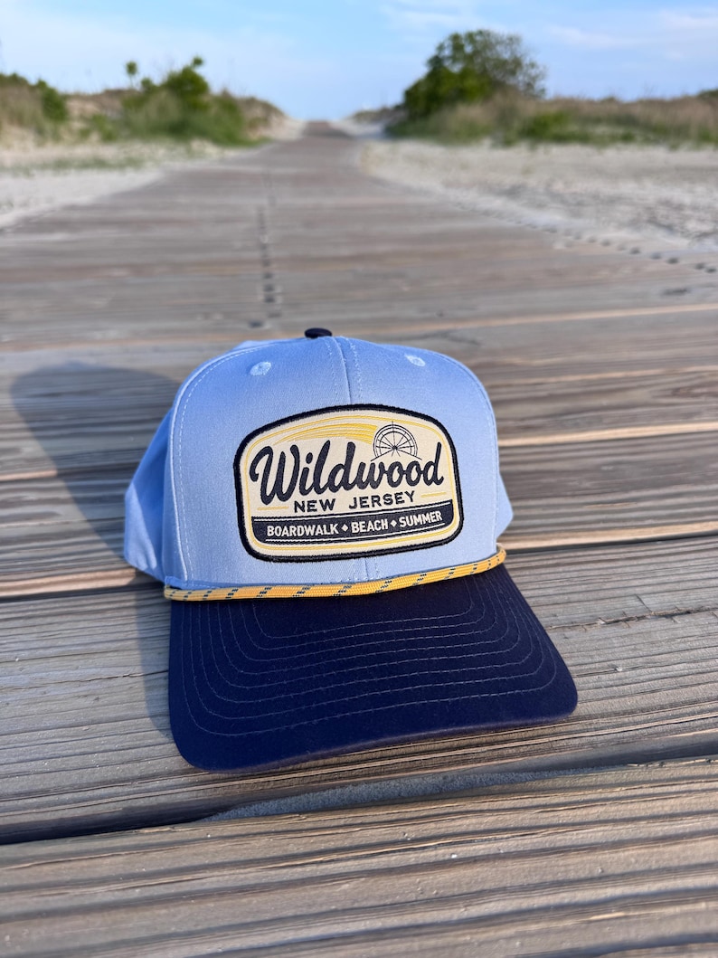 May include: A light blue baseball cap with a dark navy blue brim and a yellow rope detail. The front features a patch with the text Wildwood New Jersey Boardwalk Beach Summer and a graphic of a Ferris wheel in black, yellow, and white.