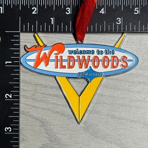 May include: A metal ornament shaped like a surfboard with the words "Welcome to the Wildwoods, New Jersey" on it. The surfboard is blue, white, and red with a yellow and silver V shape behind it.