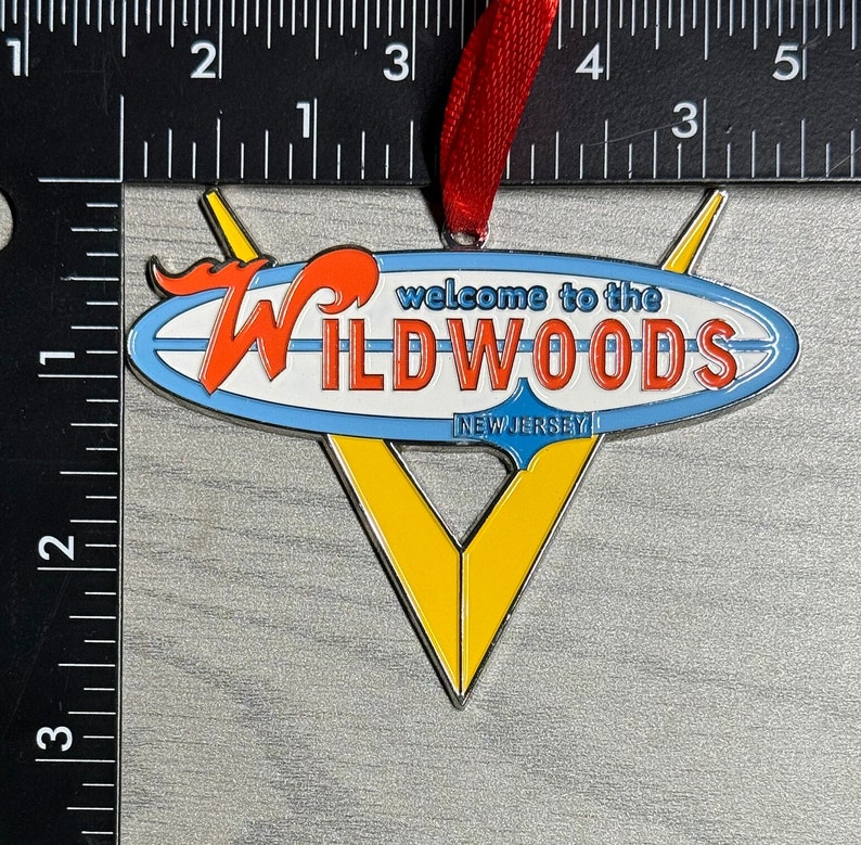 May include: A metal ornament shaped like a surfboard with the words "Welcome to the Wildwoods, New Jersey" on it. The surfboard is blue, white, and red with a yellow and silver V shape behind it.
