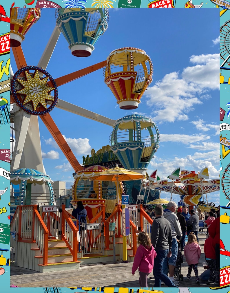 May include: A colorful Ferris wheel with gondolas shaped like hot air balloons. The Ferris wheel is on a boardwalk with people walking by. The Ferris wheel has a sign that says "Moby Dick".