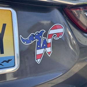 May include: A car sticker featuring a stylized letter "W" with a patriotic design. The top portion is blue with white stars, and the bottom is red and white stripes, resembling the American flag. The sticker is affixed to a gray car.
