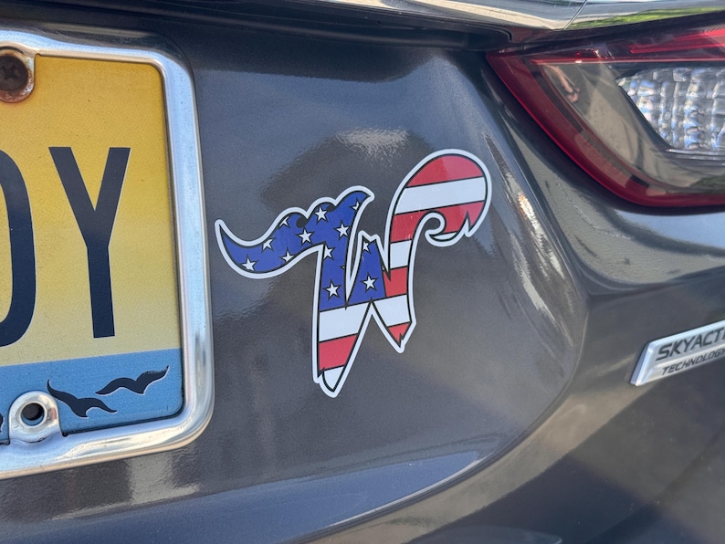May include: A car sticker featuring a stylized letter "W" with a patriotic design. The top portion is blue with white stars, and the bottom is red and white stripes, resembling the American flag. The sticker is affixed to a gray car.
