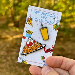 K&ouml;nnte beinhalten: Ein Set aus vier Emaille-Pins mit Motiven aus Wildwood, New Jersey. Die Pins zeigen ein St&uuml;ck Pizza, ein Glas Orangensoda, einen Teller Pommes und eine Krabbe mit Sonnenbrille. Die Pins befinden sich auf einer wei&szlig;en Karte mit dem Text "Wildwood Pin Collection" und der Website "wildwoodvideoarchive.com".