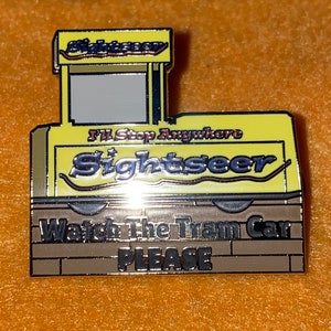 Puede incluir: Un pin de esmalte amarillo y marr&oacute;n con forma de tranv&iacute;a. El pin presenta la palabra "Sightseer" en letras plateadas, con el texto "I'll Stop Anywhere" encima. La parte inferior del pin dice "Watch The Tram Car PLEASE".