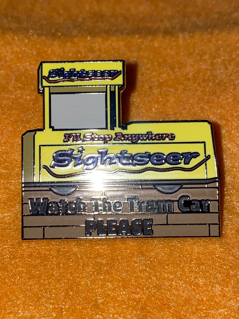 Puede incluir: Un pin de esmalte amarillo y marr&oacute;n con forma de tranv&iacute;a. El pin presenta la palabra "Sightseer" en letras plateadas, con el texto "I'll Stop Anywhere" encima. La parte inferior del pin dice "Watch The Tram Car PLEASE".